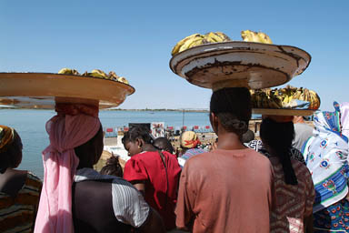 3 girls carrying bananas on their heads are watching thje boat race.