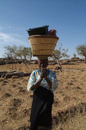 Sanga. Dogon woman front, basket on head, smiles.