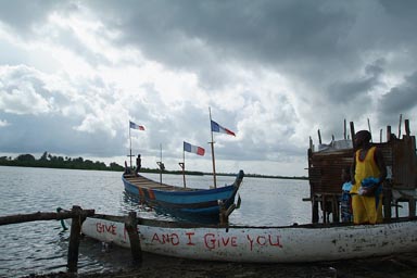 Harper, Liberia, African man in pirogue, another pirogue with many Ivorian flags. Cloudy,