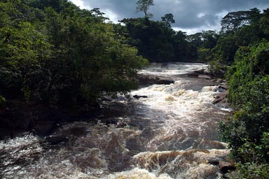 Huge brown stream, from Liberian jungle, sunlight reflects in water, dark clouds in back, Zwedru to Harper.