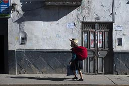 Woman and red pack walk by in morning sun, in front of white house and wooden door, Cajamarca, Peru.