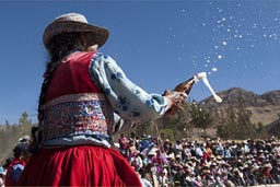 Tradition has it to offer beer, splash it around on the dust of the bull fighting arena, or better hit the bull. Old woman in red traditional bollera and hat splashes beer. Huambo, Arequipa, Peru.